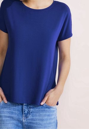 Persoon draagt een effen blauwe T-shirt met korte mouwen, lichtjes ingestopt in lichtblauwe spijkerbroek, handen in de zakken, neutrale achtergrond.