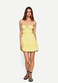 Mini abito giallo con spalline sottili, dettaglio con nodo frontale e design a strati con volant. Indossato con sandali neri con lacci. Tessuto liscio.