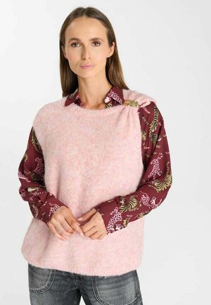 Vrouw draagt een lichtroze pluizige mouwloze trui over een bordeauxrode blouse met dierenprints, staand met handen voor zich gevouwen.