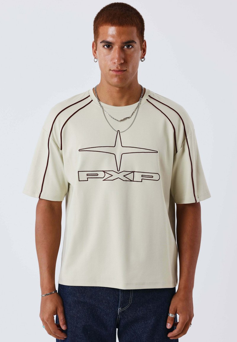 Jeune homme aux cheveux courts portant un t-shirt beige avec le logo PXP, un jean foncé, des colliers argentés, des boucles d'oreilles créoles et des bagues aux deux mains.