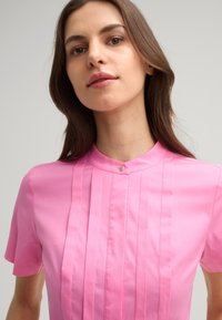 Kurzärmlige pinke Bluse mit Stehkragen, verziert mit vertikalen Falten auf der Vorderseite und einem kleinen Knopf am Ausschnitt. Glatte Textur.