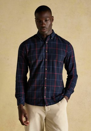 Marineblaues Karoshirt mit roten und grünen Streifen, Knopfdowkragen, langen Ärmeln und vordere Knopfverschluss. Getragen mit beigen Hosen.