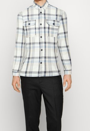 Chemise à manches longues avec boutons, motif à carreaux bleu clair, gris et blanc, poches poitrine, portée avec un pantalon noir.