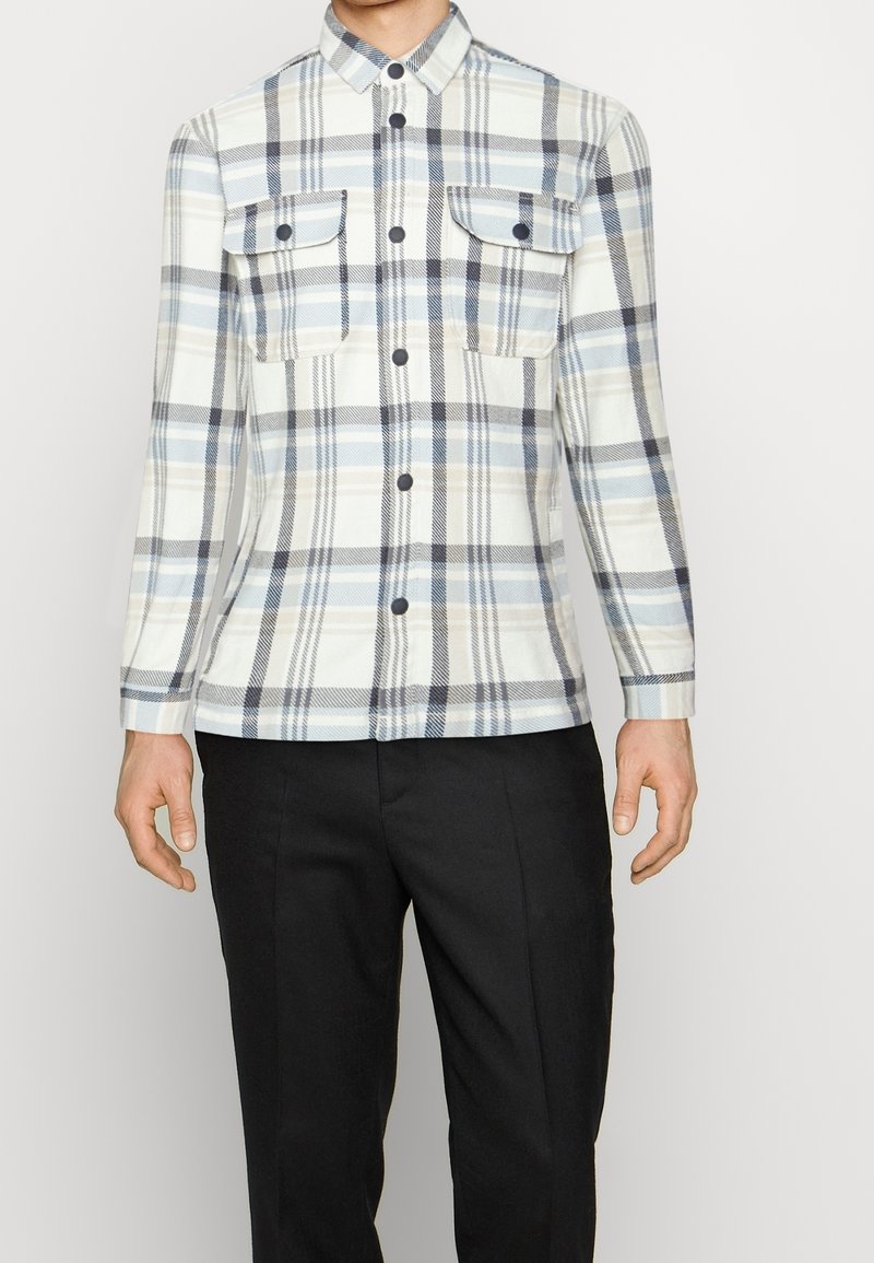 Chemise à manches longues avec boutons, motif à carreaux bleu clair, gris et blanc, poches poitrine, portée avec un pantalon noir.