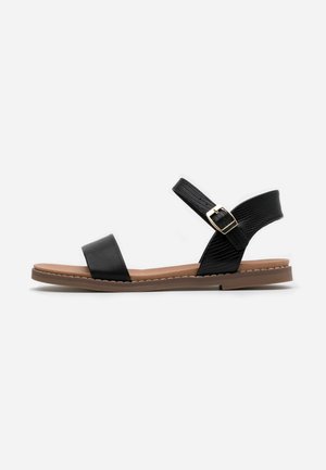 Black sandal featuring a smooth leather strap and a textured ankle band secured with a gold buckle, set on a flat brown sole.