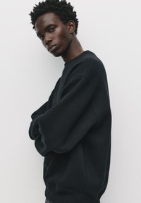 Schwarzer oversized Sweatshirt mit Rundhalsausschnitt, gerippten Bündchen und glatter Textur. Modell posiert mit verschränkten Armen vor einem weißen Hintergrund.
