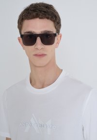 Gafas de sol rectangulares negras con sutiles acentos plateados, combinadas con una camiseta blanca simple que presenta texto de logotipo en relieve. Texturas suaves.