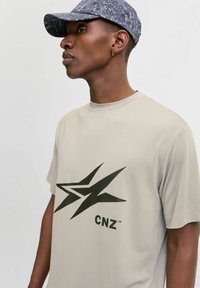 Lys beige t-shirt med korte ærmer, prydet med en stor sort stjerne grafik og teksten "CNZ" nedenunder. En struktureret mørk kasket fuldender looket.