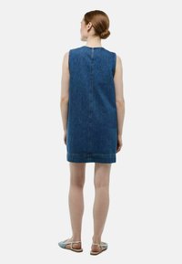 Robe en denim à coupe droite, sans manches, de couleur bleu foncé, avec des coutures visibles. Portée avec des sandales bleu clair.