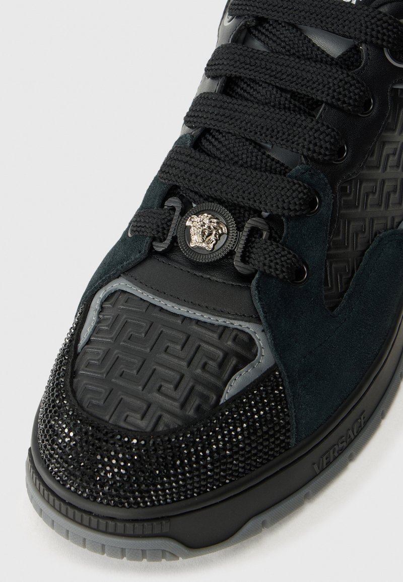 Chaussure en cuir et suède noir avec des accents métalliques, des motifs texturés et des strass sur la zone des orteils, dotée d'une semelle texturée.