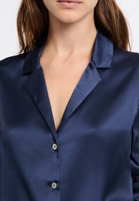 Blusa de satén azul marino con cuello con muesca, presenta un cierre frontal con botones y un brillo suave. Las mangas tienen un ajuste relajado, realzando la caída general.