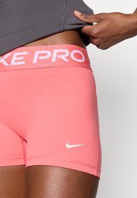 Σορτς Nike Pro σε κοραλί ροζ, με φαρδιά ελαστική ζώνη μέσης που φέρει το "NIKE PRO" σε ανοιχτό ροζ και μικρό λευκό λογότυπο στο πόδι.