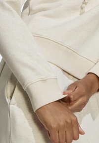 Beige sweatshirt med ribbade manschetter, med en mjuk textur och synlig sömnad. Händer som drar upp manschetten något.