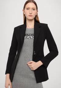 Blazer nero su misura con rever affilati, chiusura a un bottone e tasche laterali. Indossato sopra un abito a maglia grigio a strisce con dettaglio del logo.
