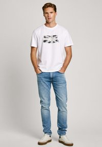 Biała bawełniana koszulka z grafiką flagi Wielkiej Brytanii, zestawiona z jasnoniebieskimi dżinsami i białymi trampkami z brązowymi akcentami.