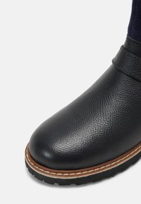 Czarny skórzany botek do kostki o teksturowanym wykończeniu, zaokrąglonym nosku oraz kontrastowej brązowej podeszwie z białym przeszyciem. Minimalistyczny design.