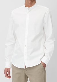 Vit skjorta med krage, tillverkad av mjukt bomullstyg. Har långa ärmar och en diskret röd accent vid fållen.