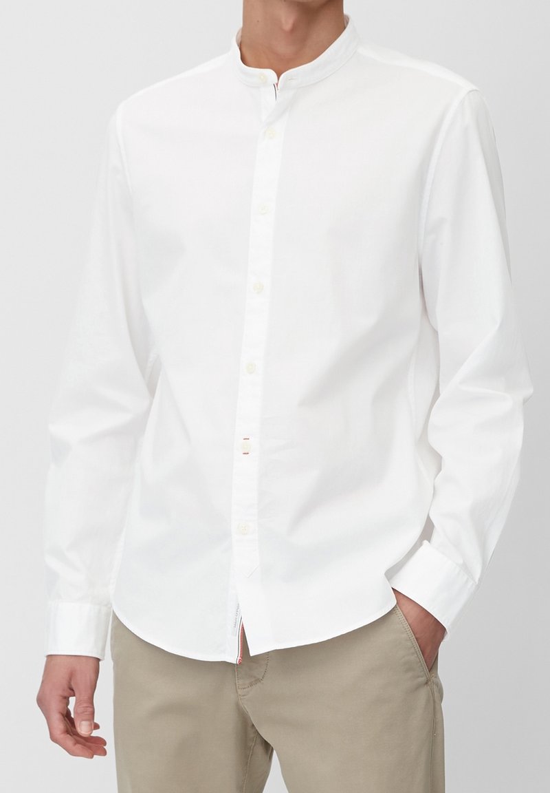 Vit skjorta med krage, tillverkad av mjukt bomullstyg. Har långa ärmar och en diskret röd accent vid fållen.