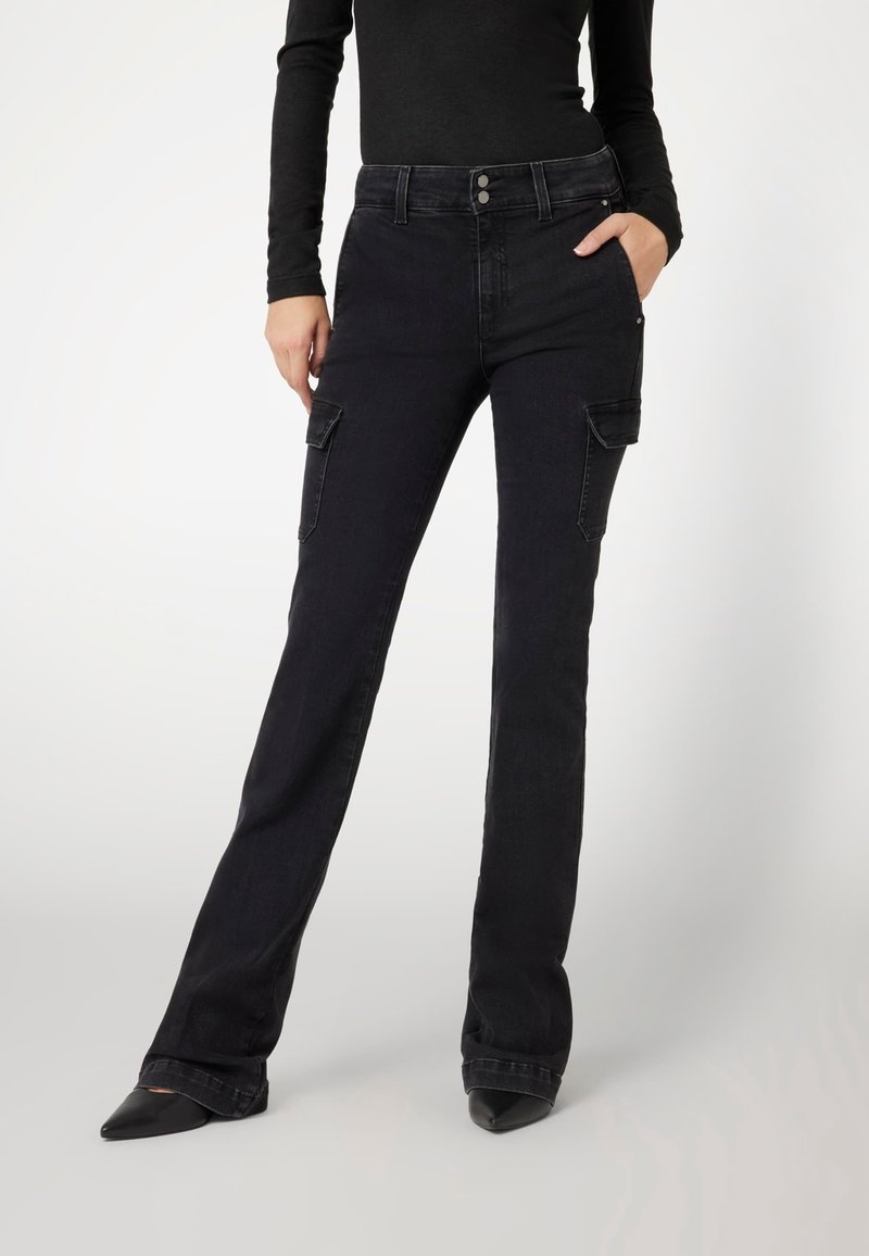 Femme portant un jean noir taille haute, coupe droite, avec des poches cargo et des chaussures noires à bouts pointus, main dans la poche, debout sur fond blanc.