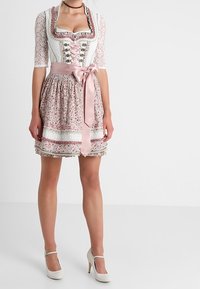 Femme portant une robe traditionnelle dirndl rose et blanche avec des manches en dentelle, un nœud en satin à la taille et des chaussures à talons hauts blanches.