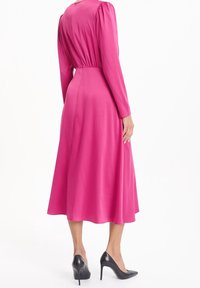 Robe rose à manches longues avec une taille froncée, une jupe fluide et une texture lisse et brillante. Portée avec des chaussures à talons noirs.