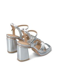 Stříbrné sandály na podpatku s robustním blokovým podpatkem, několika křížovými pásky a detailem kulaté spony, s hladkým metalickým povrchem a béžovou podrážkou.