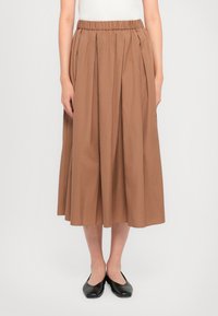 Brun, kneelengde skjørt laget av bomull med elastisk midjebånd, med knivfeller og en jevn tekstur. Båret med svarte ballerinas.