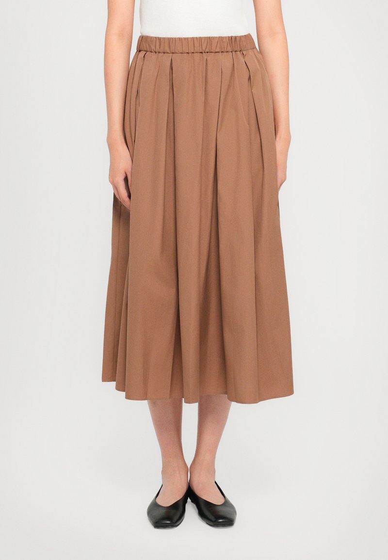 Brun, kneelengde skjørt laget av bomull med elastisk midjebånd, med knivfeller og en jevn tekstur. Båret med svarte ballerinas.