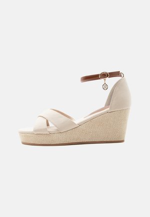 Beige kile-sandal med teksturert platå, kryssede stoffremmer og brun ankelstropp med liten rund gullsmykke.