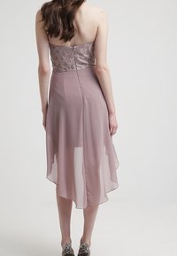 Šaty bez ramínek s texturovaným krajkovým živůtkem v světle fialové barvě, splývavou sukní z chiffonového materiálu s asymetrickým lemem a jemným leskem.