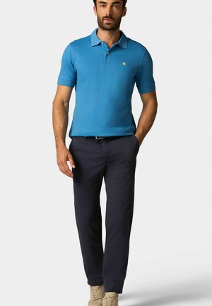 Tyrkysové polo tričko s krátkým rukávem, se malým zeleným logem, v kombinaci s tmavě námořnickými chinos kalhotami a světle béžovými sneakers.