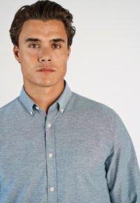 Ανοιχτό μπλε πουκάμισο με γιακά, κατασκευασμένο από υφασμάτινη ύφανση, με κρεμ κουμπιά και ορατές ραφές μπροστά και στα μανίκια.