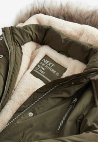 Veste matelassée vert olive avec une doublure en fausse fourrure beige. Elle présente une fermeture éclair, des boutons-pression, et une étiquette indiquant "NEXT THE FUTURE IS NOW."