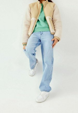 Personne portant une veste en polaire beige, un sweat-shirt vert, un jean bleu clair et des baskets blanches, debout avec une jambe levée sur un fond blanc.