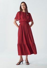 Crvena maksi haljina s kratkim rukavima, smotanim strukom i slojevitom donjištem. Lagani materijal s glatkom teksturom. Uparena s crnim sandalama na visoku petu.