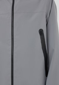 Giacca leggera grigia con una texture liscia, dotata di una cerniera nera e di una tasca laterale con zip per una maggiore funzionalità.