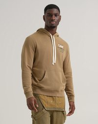 Felpa cappuccio marrone con tasca frontale, caratterizzata da un laccio bianco e dettagli con logo. Il modello indossa pantaloni a righe gialle.