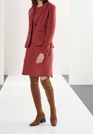 Blazer e vestito a fantasia rossa con un motivo a zigzag testurizzato. Stivali marroni alti fino alla coscia con punta quadrata e tacco spesso.