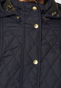 Granatowa pikowana kurtka z wysokim kołnierzem, ozdobiona złotymi zatrzaskami i kontrastową podszewką widoczną na krawędzi kołnierza.