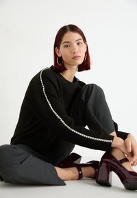 Maglione nero strutturato con dettagli a strisce bianche, abbinato a pantaloni grigi. La modella indossa scarpe col tacco lucide bordeaux.