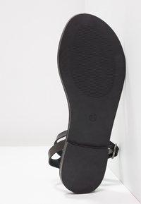 Sandale plate noire avec bride de cheville ajustable et semelle texturée, taille 37, positionnée droite contre un coin blanc.