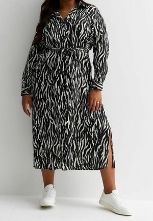 Frau trägt ein langärmliges Midikleid mit schwarz-weißem Zebramuster und seitlichem Schlitz, kombiniert mit weißen Turnschuhen, und steht vor einem weißen Hintergrund.