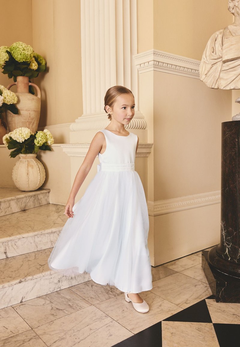 Jeune fille en robe blanche descendant un escalier en marbre dans une pièce élégante avec des colonnes classiques et des arrangements floraux.
