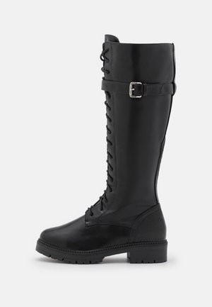 Botte montante noire en cuir avec lacets, sangle à boucle latérale, talon épais et semelle texturée, vue de profil sur fond blanc.