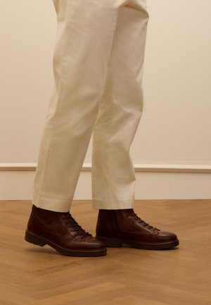 Bottes en cuir marron à lacets avec une finition texturée, semelle robuste et œillets, associées à un pantalon beige cintré sur un sol en bois.