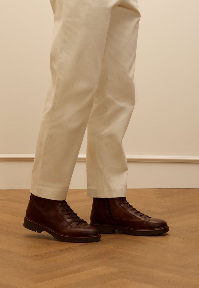 Bruine leren veterschoenen met een gestructureerde afwerking, stevige zool en gaatjes, gecombineerd met crème-kleurige tapered broek op een houten vloer.