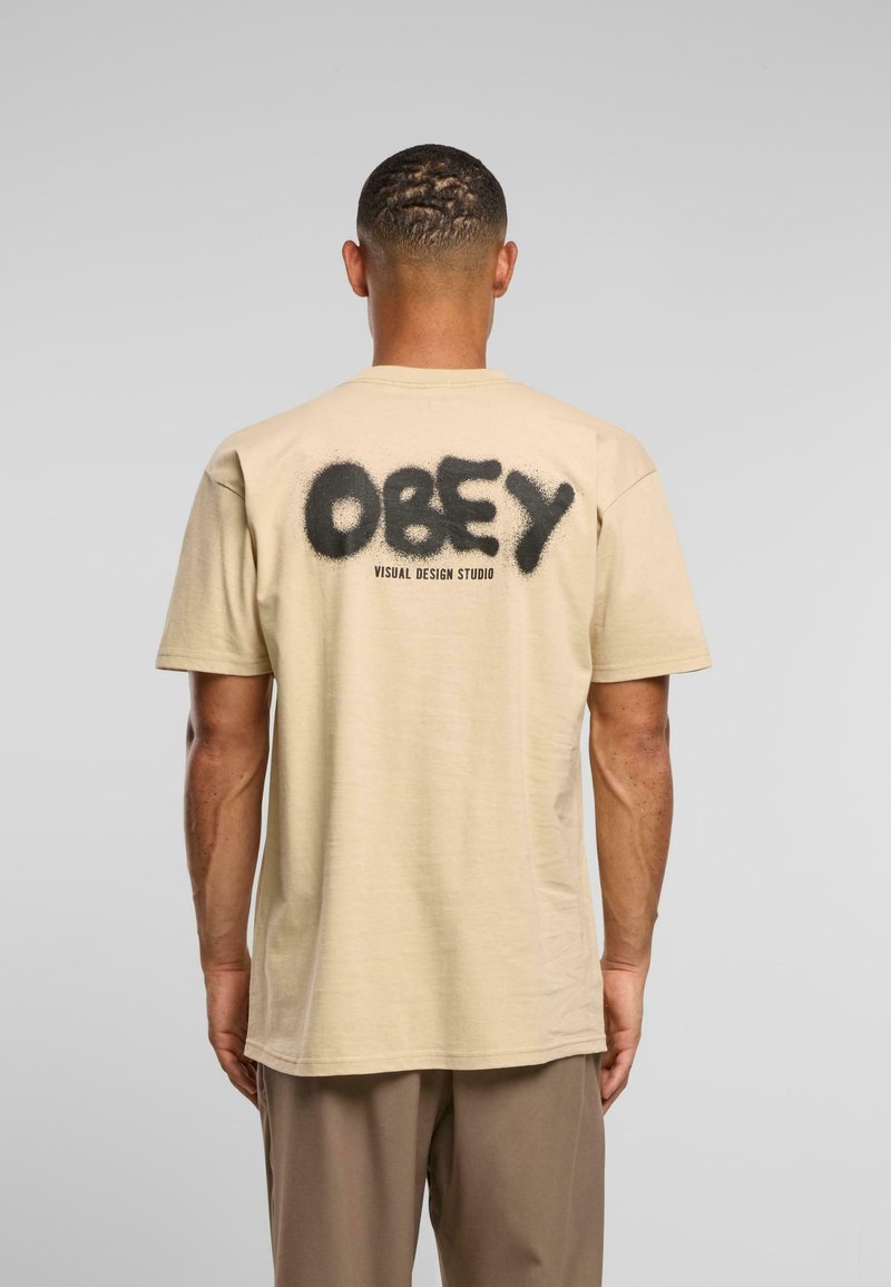Muž otočený zády, oblečený v béžovém tričku s černým textem "OBEY VISUAL DESIGN STUDIO" ve stylu sprejového nátěru na zádech a hnědých kalhotách.