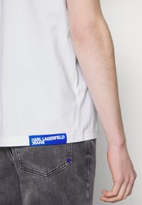 Balta T-kreklu ar zilo Karl Lagerfeld Jeans birku apakšmalā, kombinējot ar pelēkajām džinsām ar nelielu zilu akcentu pie kabatas.