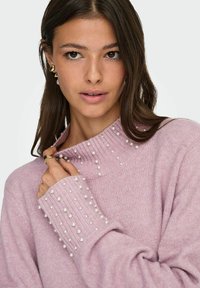 Kvinne med langt brunt hår iført en rosa genser utsmykket med små hvite perler på kragen og mansjettene, som holder kragen forsiktig.