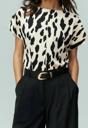 Femme portant un haut blanc à manches courtes avec un imprimé animal noir, rentré dans un pantalon noir taille haute avec une ceinture noire et une boucle dorée.